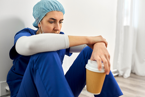 medicine, healthcare and pandemic concept - close up of sad young female doctor or nurse with disposable cup of takeaway coffee sitting on floor at hospital