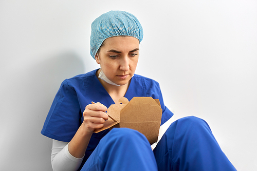 medicine, healthcare and pandemic concept - sad and tired young female doctor or nurse eating takeaway food from disposable box sitting on floor at hospital