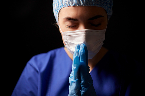 health, medicine and pandemic concept - sad young female doctor or nurse wearing face protective mask for protection from virus disease praying over black background