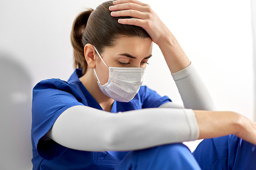 medicine, healthcare and pandemic concept - sad young female doctor or nurse wearing face protective mask for protection from virus disease sitting on floor and holding to head