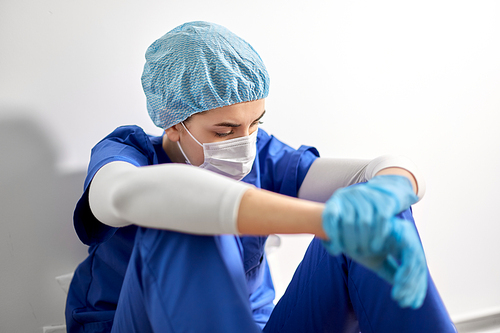 medicine, healthcare and pandemic concept - sad young female doctor or nurse wearing face protective mask for protection from virus disease sitting on floor at hospital