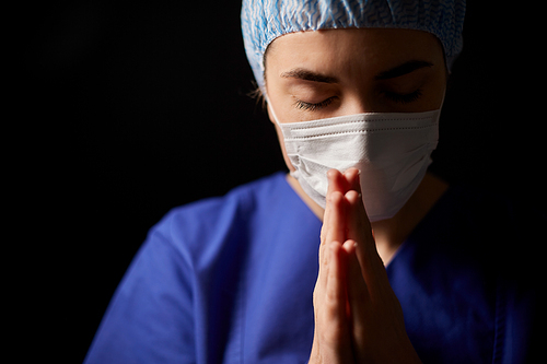 health, medicine and pandemic concept - sad young female doctor or nurse wearing face protective mask for protection from virus disease praying over black background