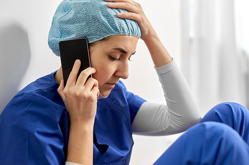 medicine, healthcare and pandemic concept - close up of sad young female doctor or nurse sitting on floor and calling on smartphone