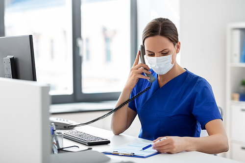 medicine, people and healthcare concept - female doctor or nurse wearing face protective medical mask for protection from virus disease with computer and clipboard calling on phone at hospital