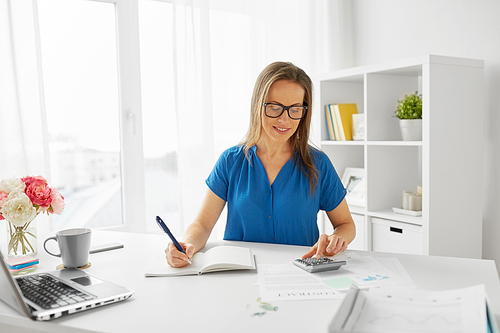 business, finance and accounting concept - middle-aged woman with calculator counting and writing to notebook at office