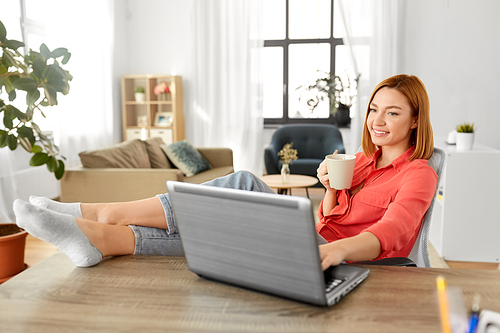 remote job, technology and people concept - happy smiling young woman with laptop computer drinking coffee at home office and resting her feet on table