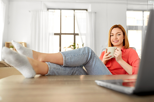 remote job, technology and people concept - happy smiling young woman with laptop computer drinking coffee at home office and resting her feet on table