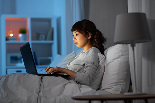 technology, internet, communication and people concept - happy smiling young asian woman with laptop computer lying in bed at home at night