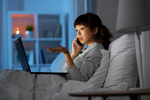 technology, internet, communication and people concept - young asian woman with laptop computer calling on smartphone in bed at home at night