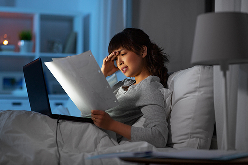 technology, remote job and people concept - stressed young asian woman with laptop computer and papers working in bed at home at night