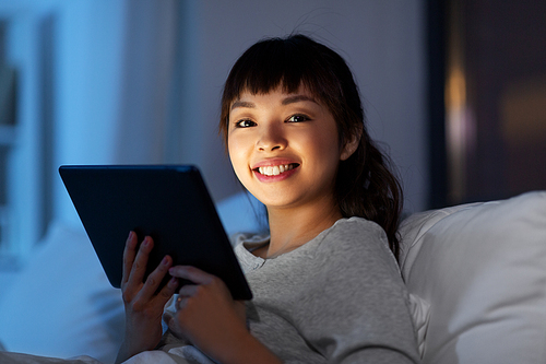 technology, internet and people concept - happy smiling young asian woman with tablet pc computer lying in bed at home at night