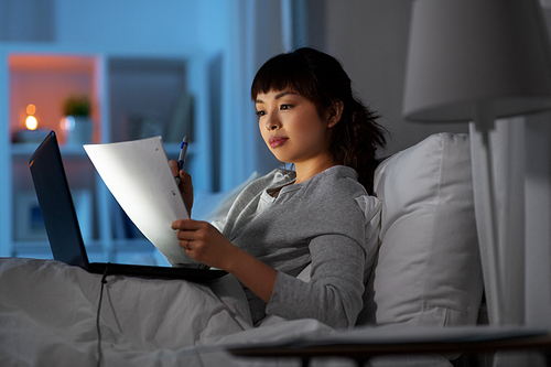 technology, remote job and people concept - young asian woman with laptop computer and papers working in bed at home at night