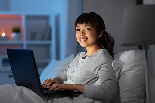 technology, internet, communication and people concept - happy smiling young asian woman with laptop computer lying in bed at home at night