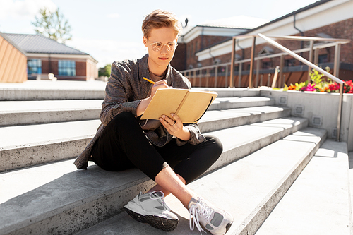 leisure, education and people concept - young man or teenage boy in glasses with notebook, diary or sketchbook writing or drawing in city