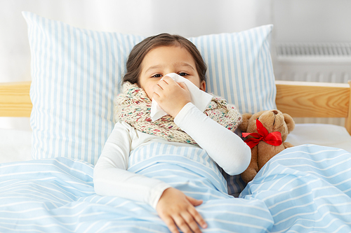 health, children and people concept - sick little girl lying in bed and blowing nose with paper tissue at home