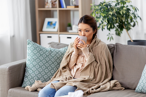 health, cold and people concept - sad sick young asian woman in blanket drinking hot tea at home