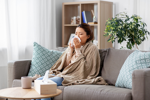 health, cold and people concept - sad sick young asian woman in blanket blowing nose with paper tissue at home