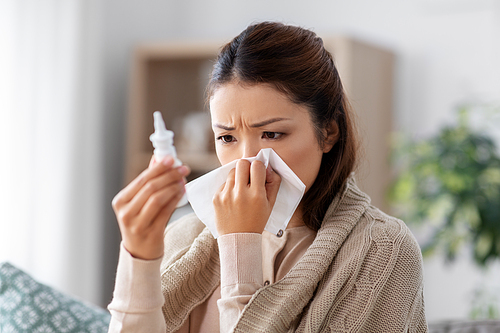 health, cold and people concept - sad sick young asian woman in blanket with nasal spray medicine and paper tissue at home