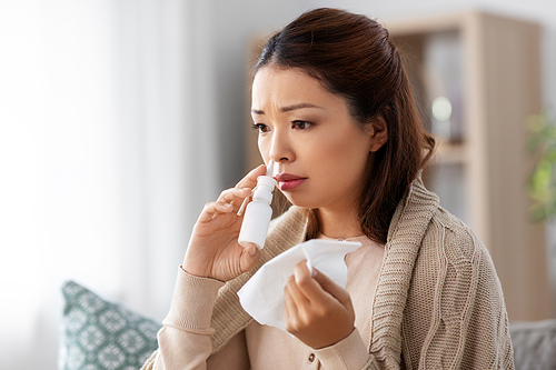 health, cold and people concept - sad sick young asian woman in blanket using nasal spray medicine and paper tissue at home