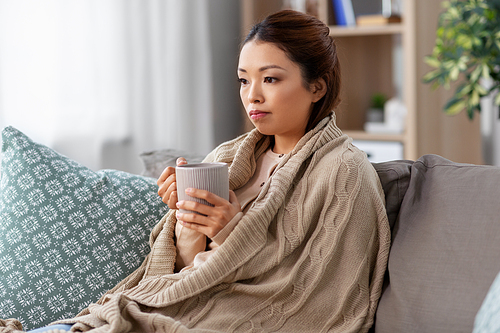 health, cold and people concept - sad sick young asian woman in blanket drinking hot tea at home