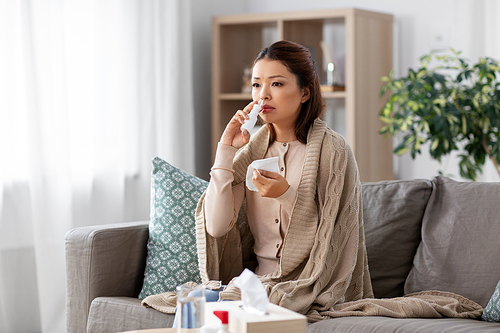 health, cold and people concept - sad sick young asian woman in blanket using nasal spray medicine and paper tissue at home