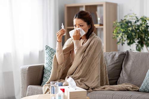 health, cold and people concept - sad sick young asian woman in blanket with nasal spray medicine and paper tissue at home