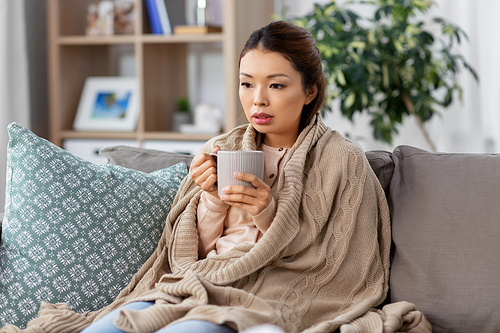health, cold and people concept - sad sick young asian woman in blanket drinking hot tea at home