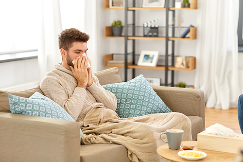 healthcare, cold, hygiene and people concept - sick man in blanket with paper tissue blowing his nose at home