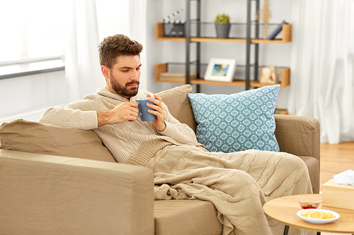 health, cold and people concept - sick young man in blanket drinking hot tea with lemon and honey at home