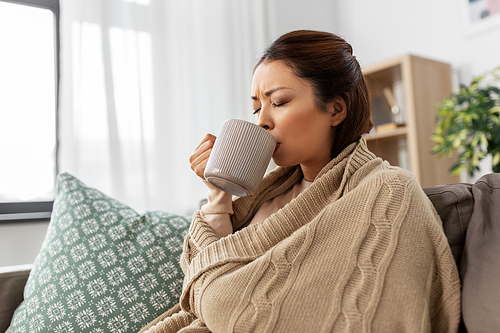 health, cold and people concept - sick young asian woman in blanket drinking hot tea at home