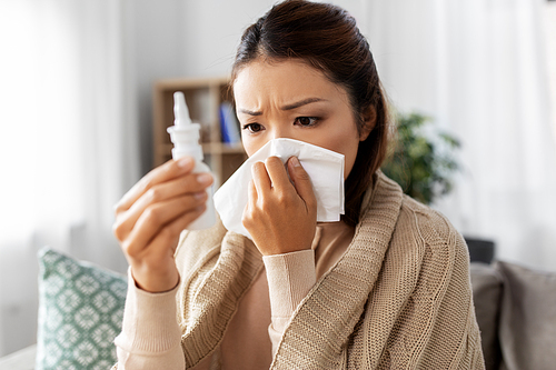 health, cold and people concept - sick young asian woman in blanket with nasal spray medicine blowing nose to paper tissue at home