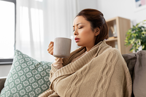 health, cold and people concept - sick young asian woman in blanket drinking hot tea at home