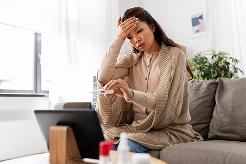 healthcare, technology and people concept - sick asian woman in blanket with thermometer having video call on tablet pc computer at home