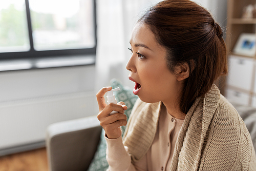 health, cold and people concept - sad sick young asian woman in blanket using oral spray medicine at home