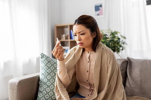 health, cold and people concept - sad sick young asian woman in blanket with nasal spray medicine at home