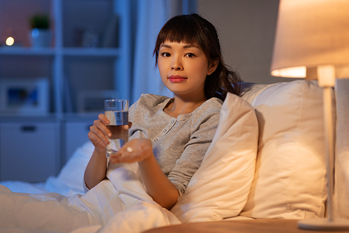 people, health and bedtime concept - stressed asian american woman having headache and taking medicine or sleeping pill with water in bed at night