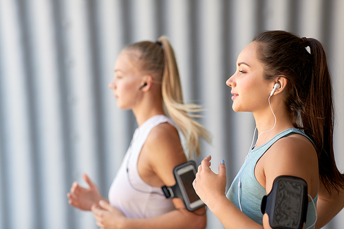 fitness, sport and healthy lifestyle concept - young women or female friends with earphones wearing armbands with smartphones and running outdoors