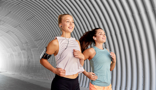 fitness, sport and healthy lifestyle concept - young women or female friends with earphones wearing armbands with smartphones and running outdoors