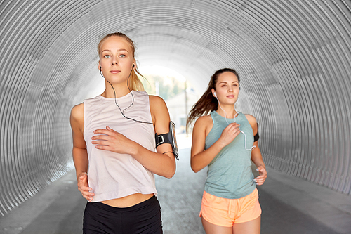 fitness, sport and healthy lifestyle concept - young women or female friends with earphones wearing armbands with smartphones and running outdoors