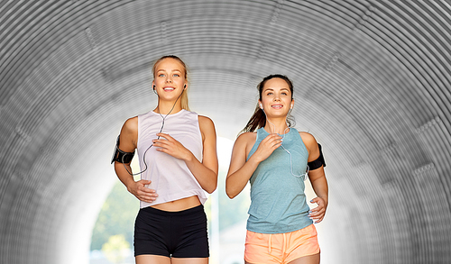 fitness, sport and healthy lifestyle concept - happy young women or female friends with earphones wearing armbands with smartphones and running outdoors