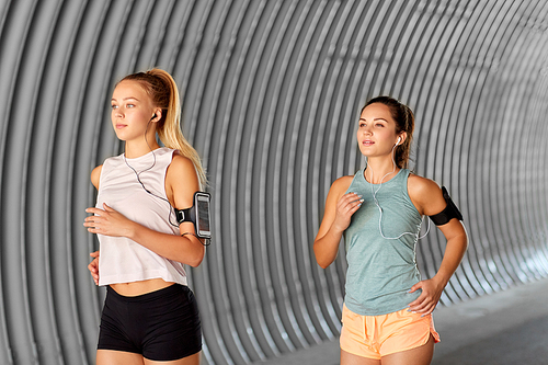 fitness, sport and healthy lifestyle concept - young women or female friends with earphones wearing armbands with smartphones and running outdoors