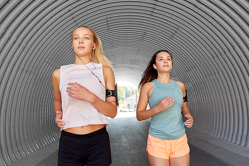 fitness, sport and healthy lifestyle concept - young women or female friends with earphones wearing armbands with smartphones and running outdoors