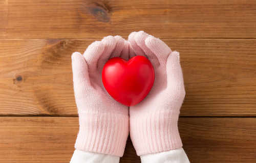 winter, valentine's day and christmas concept - hands in pale pink woollen gloves holding red heart over wooden boards background