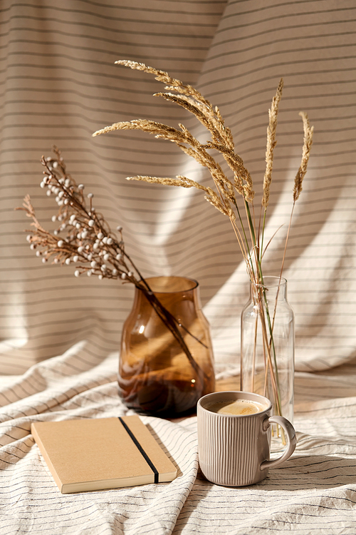 still life, hygge and drinks concept - cup of coffee, diary, decorative dried flowers in vase and bottle with drapery