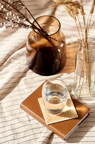 home improvement and decoration concept - still life of water in glass, book, decorative dried flowers in vase and bottle over drapery