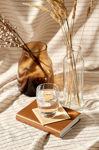 home improvement and decoration concept - still life of water in glass, book, decorative dried flowers in vase and bottle over drapery