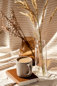 still life, hygge and drinks concept - cup of coffee on book, decorative dried flowers in vase and bottle with drapery