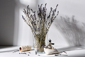 aromatherapy and organic concept - still life of dried lavender flowers in glass vase, craft sachet bag and rope dropping shadows on white surface
