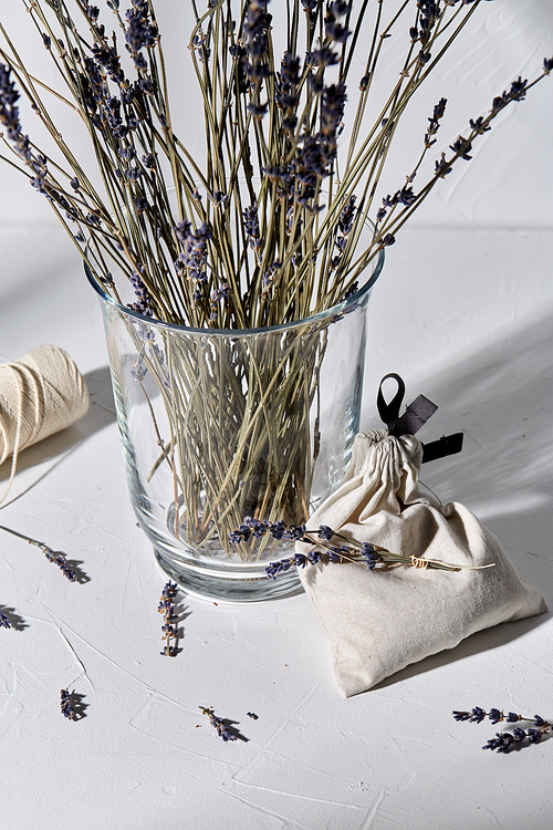 aromatherapy and organic concept - still life of dried lavender flowers in glass vase, craft sachet bag and rope dropping shadows on white surface