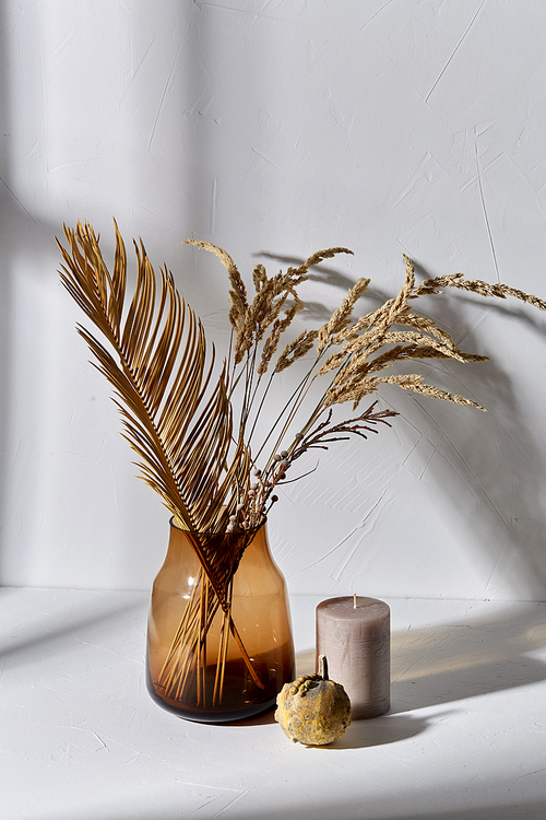 home improvement and decoration concept - still life of decorative dried flowers in brown glass vase, candle and pumpkin dropping shadows on white surface